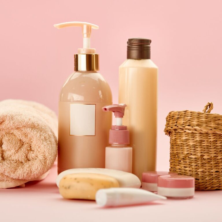 Pompflessen met crème en lotion, een stuk zeep, kleine potjes en een opgerolde handdoek op een roze achtergrond
