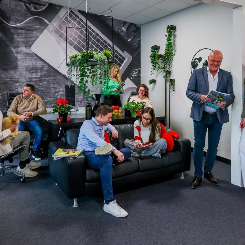 Groep collega's in een kantoorruimte met bank, planten en een grote foto van een laptop aan de muur