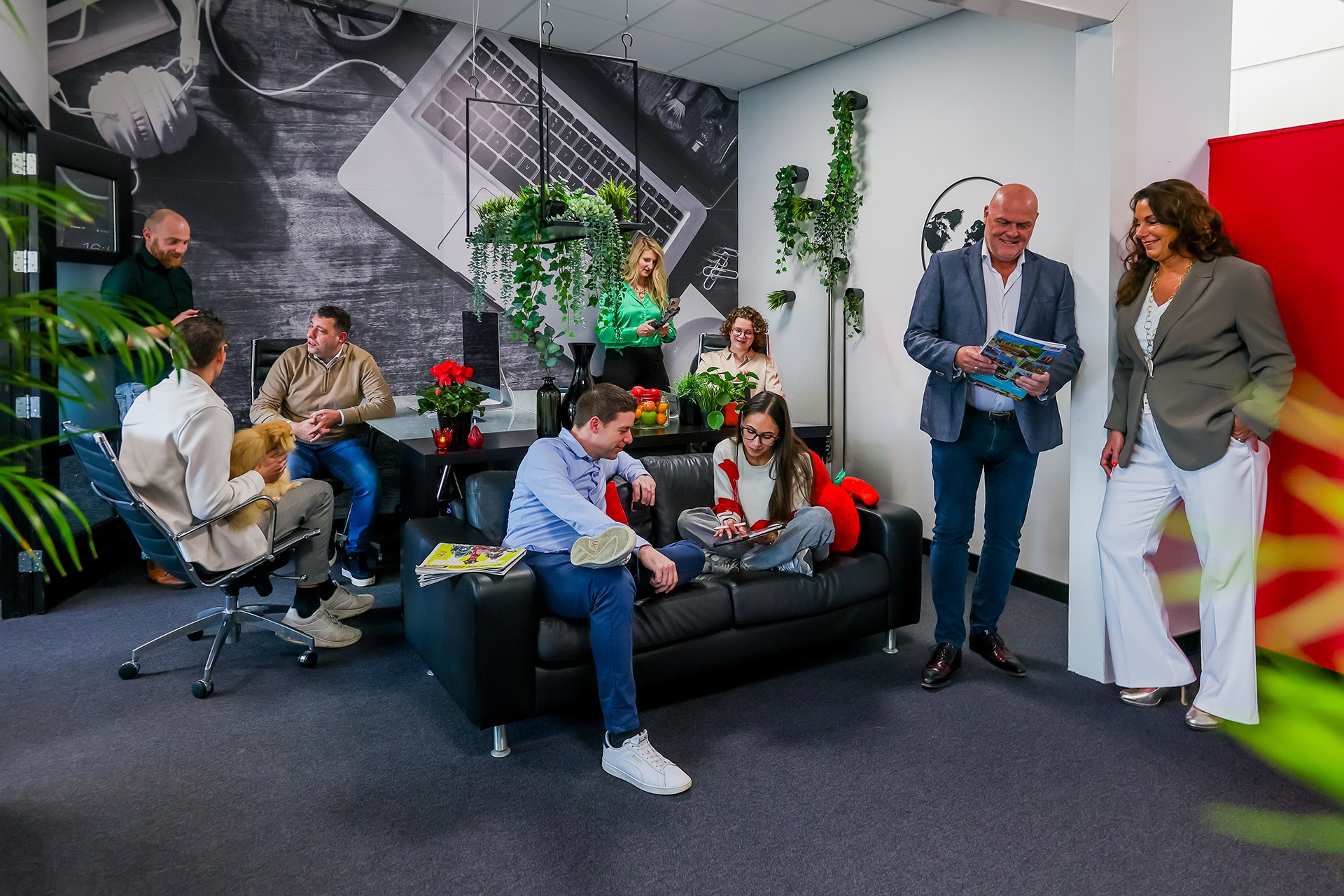 Groep collega's in een kantoorruimte met bank, planten en een grote foto van een laptop aan de muur