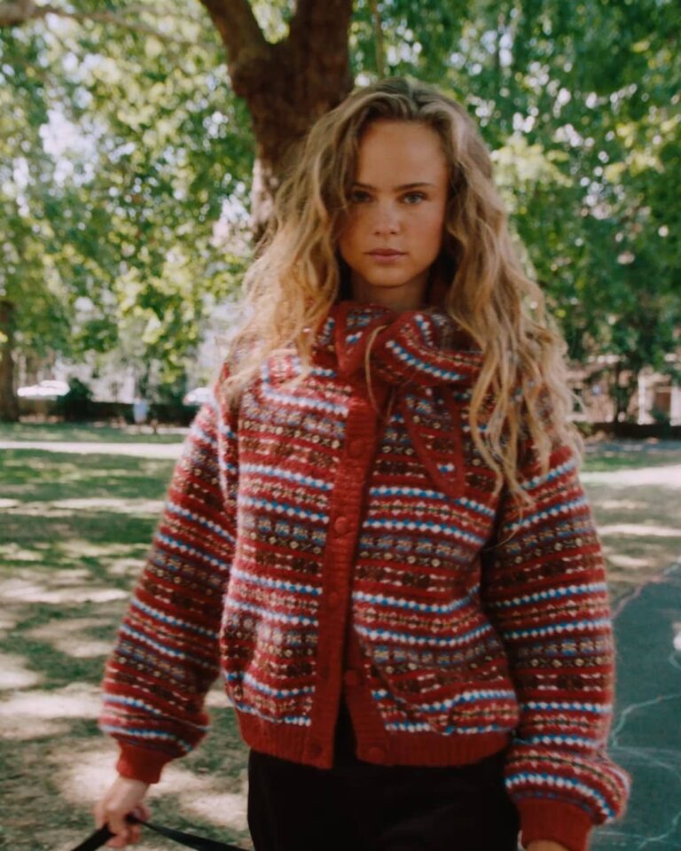 Vrouw in een rood gebreid vest met streeppatroon loopt door een park
