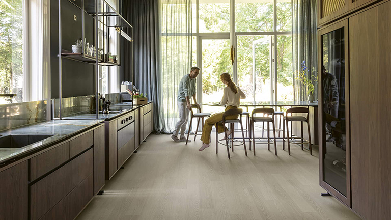 Een stel aan de bar in een lichte open keuken met een brede houtlook vloer van wand tot raam, vloeren inkopen via Grootmarkt