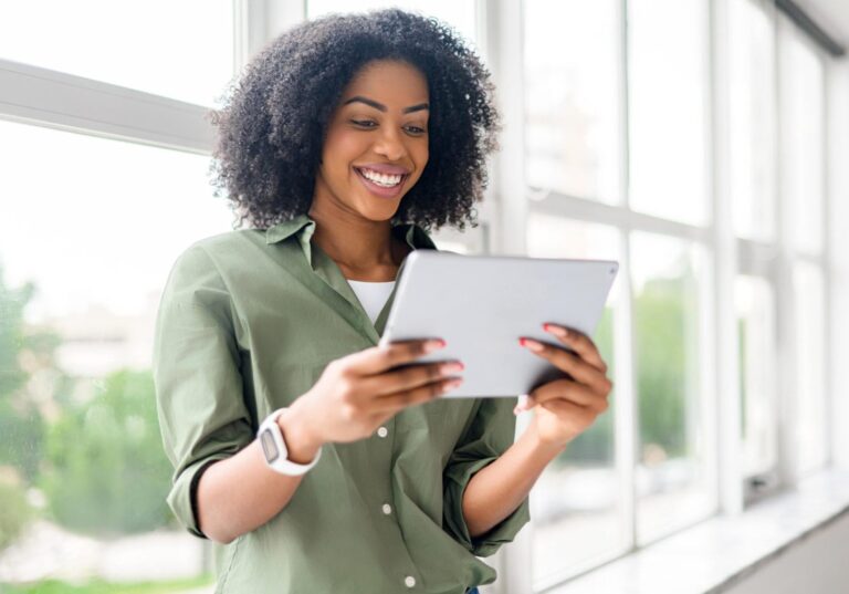 Een lachende vrouw in een groene blouse kijkt bij een raam op een tablet