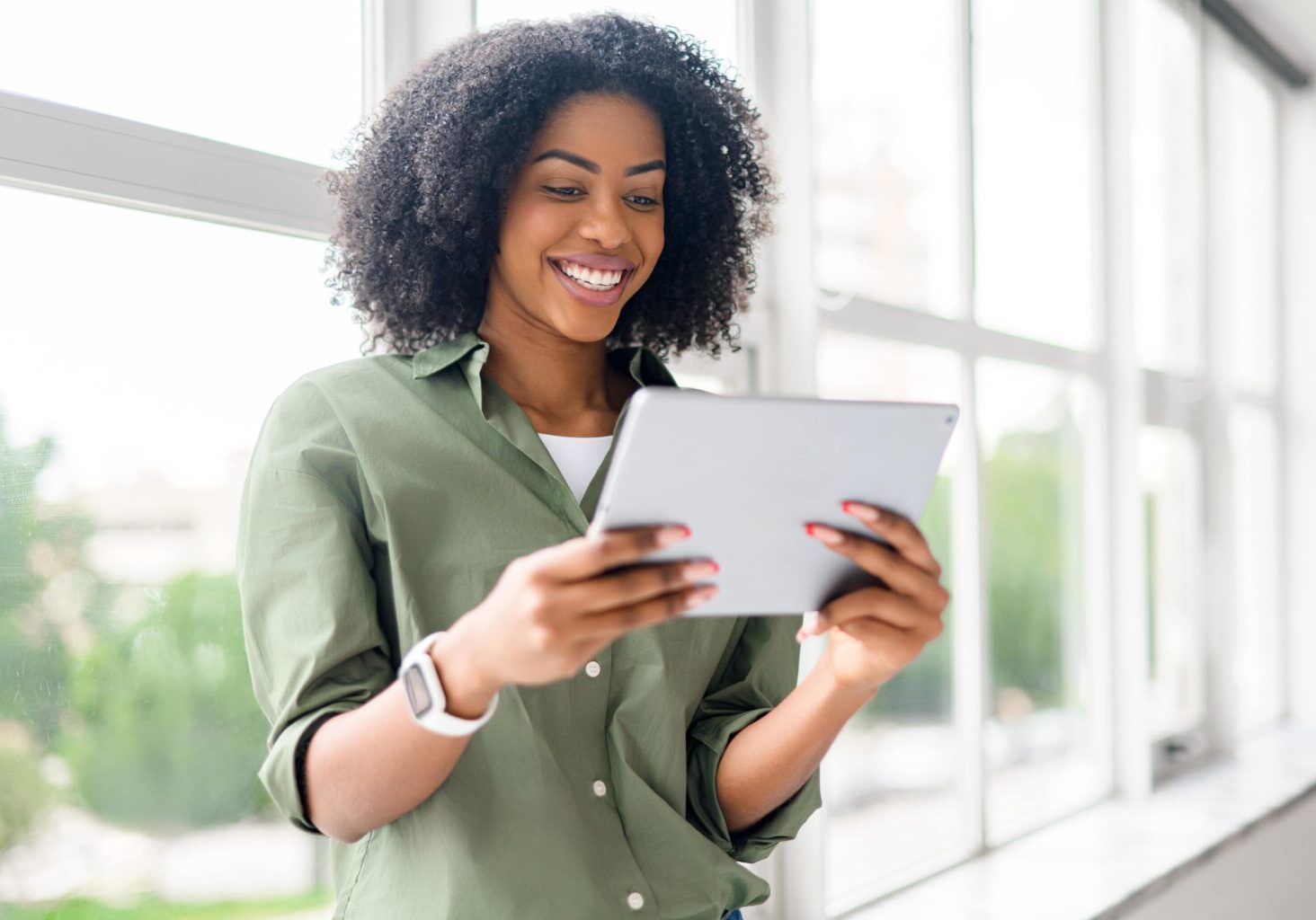 Een lachende vrouw in een groene blouse kijkt bij een raam op een tablet