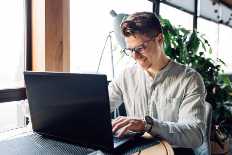 Een lachende man met bril typt op een laptop aan een tafel bij het raam