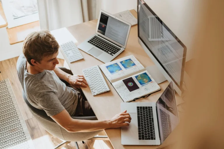 Een man aan een bureau met twee laptops, een groot scherm en een opengeslagen schrift