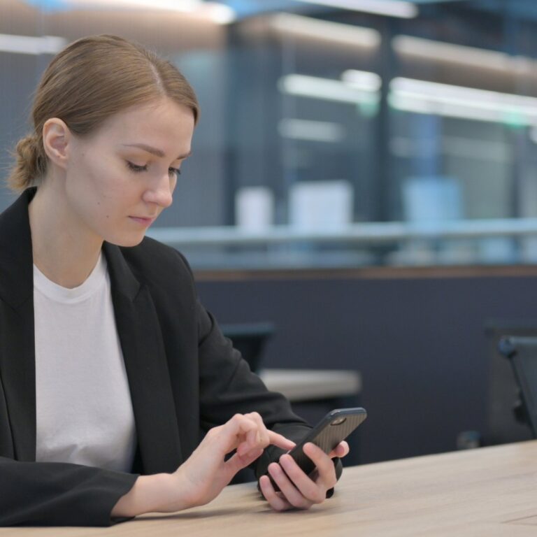Een vrouw in een zwart colbert kijkt aan tafel op haar telefoon