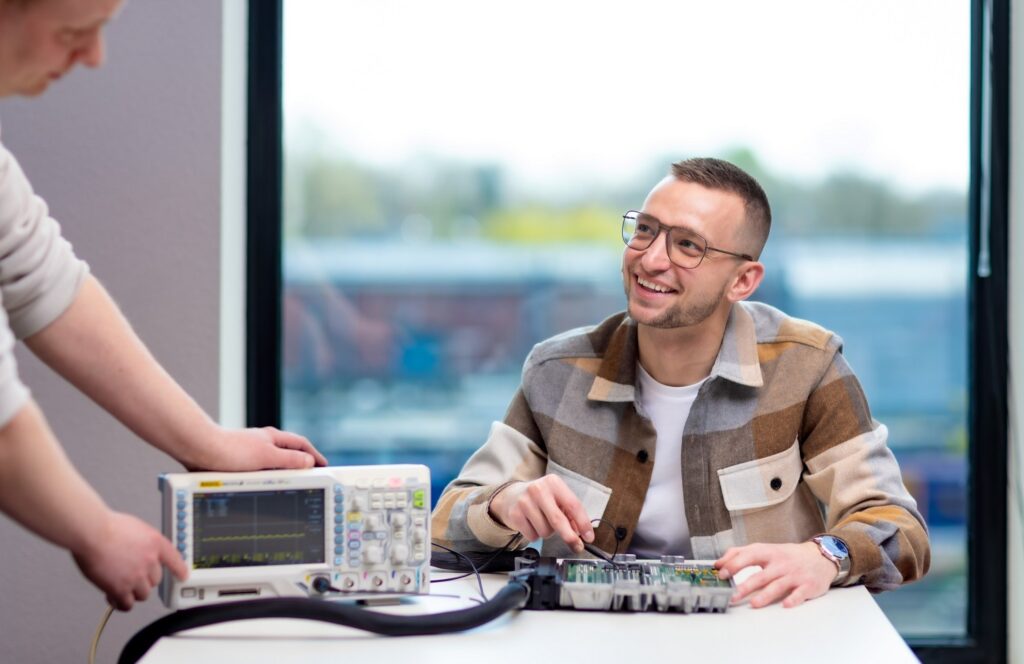 Lachende technicus test een printplaat met een oscilloscoop in een lichte werkruimte, elektronica inkopen via Grootmarkt