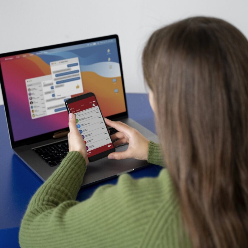 Vrouw in een groene trui kijkt op haar smartphone voor een laptop