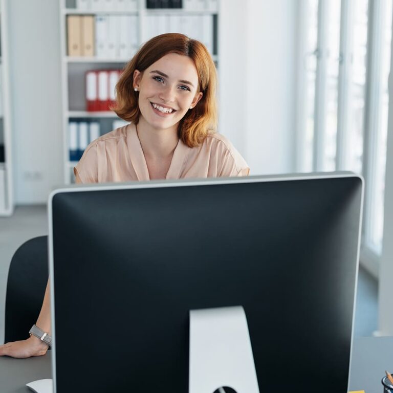 Een lachende vrouw achter een computerscherm in een licht kantoor met ordners